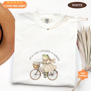 May include: White t-shirt featuring a frog in a dress and scarf riding a bicycle, with a basket of flowers. The text reads "off to cause a Kerfuffle at mahjong." The shirt also has the text "I LOVE THIS SHIRT" and "WHITE".