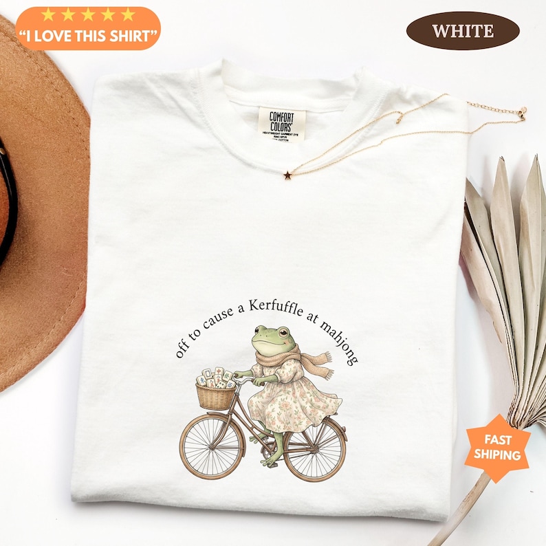 May include: White t-shirt featuring a frog in a dress and scarf riding a bicycle, with a basket of flowers. The text reads "off to cause a Kerfuffle at mahjong." The shirt also has the text "I LOVE THIS SHIRT" and "WHITE".
