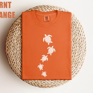 May include: Burnt orange t-shirt with a crew neck. The shirt features a design of five white sea turtles swimming downwards. The words "Burnt Orange" are printed in the upper left corner.