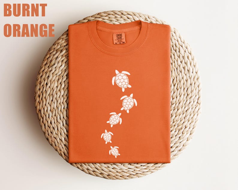 May include: Burnt orange t-shirt with a crew neck. The shirt features a design of five white sea turtles swimming downwards. The words "Burnt Orange" are printed in the upper left corner.