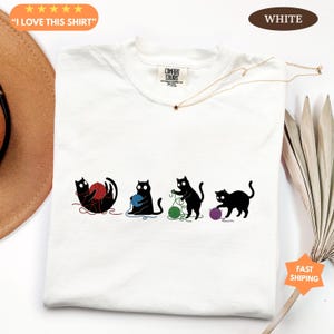 May include: White Comfort Colors t-shirt with a graphic of four black cats playing with yarn in red, blue, green, and purple. The text "I LOVE THIS SHIRT" is in an orange oval. The shirt is labeled "WHITE".