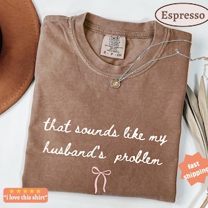 May include: Espresso-colored t-shirt with the phrase "that sounds like my husband's problem" in white script. A pink bow graphic is below the text. A silver necklace with a pendant is on the shirt.