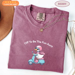 May include: Berry-coloured t-shirt featuring the phrase "Off To Be The Fun Aunt" and a cartoon duck on a blue scooter. The duck is wearing a pink helmet, sunglasses, and boots. A gold necklace is draped across the shirt.