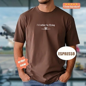 May include: A brown t-shirt with the phrase "i'd rather be flying" and a small aeroplane graphic. The shirt is espresso-coloured, with a crew neck and short sleeves. A sticker says "I love this shirt".