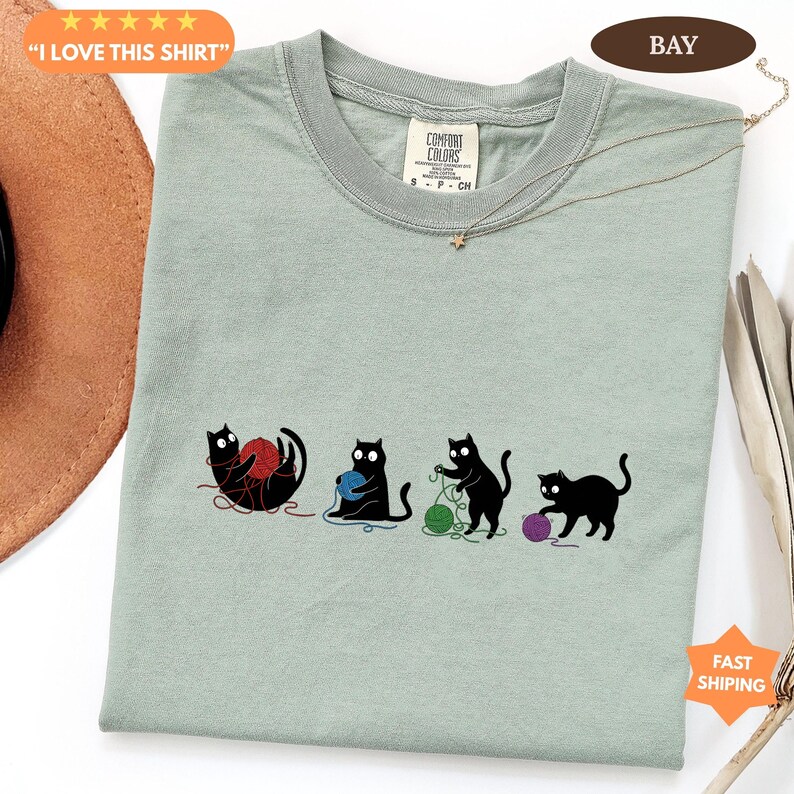 May include: Sage green t-shirt with a playful graphic of four black cats interacting with yarn balls in red, blue, green, and purple. The shirt has the text "I LOVE THIS SHIRT" and a tag that says "Comfort Colors".