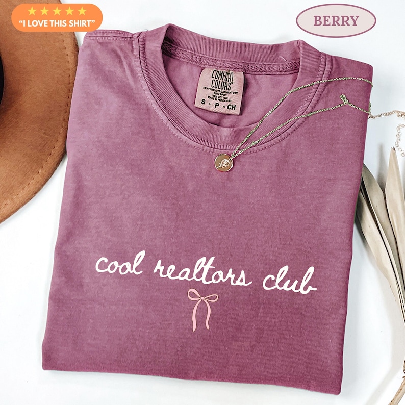 May include: A berry-colored short-sleeve t-shirt with a crew neckline. The shirt features the white text "cool realtors club" printed on the front, accompanied by a small white bow graphic below the text. A gold-toned necklace rests on the shirt.