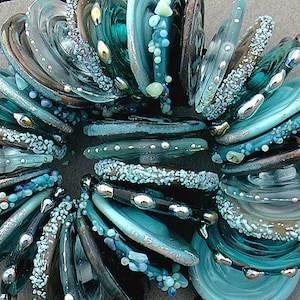 May include: A close-up of a collection of teal and silver glass beads. The beads are round and have a textured surface. They are arranged in a circular pattern.