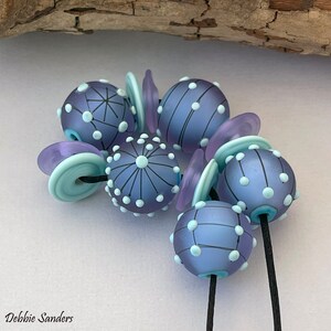 Pode incluir: Close-up de contas de vidro artesanais em tons de roxo e azul. As contas s&atilde;o decoradas com pontos brancos e linhas pretas. Algumas contas s&atilde;o redondas, outras planas ou cil&iacute;ndricas. As contas s&atilde;o enfiadas em um cord&atilde;o preto.