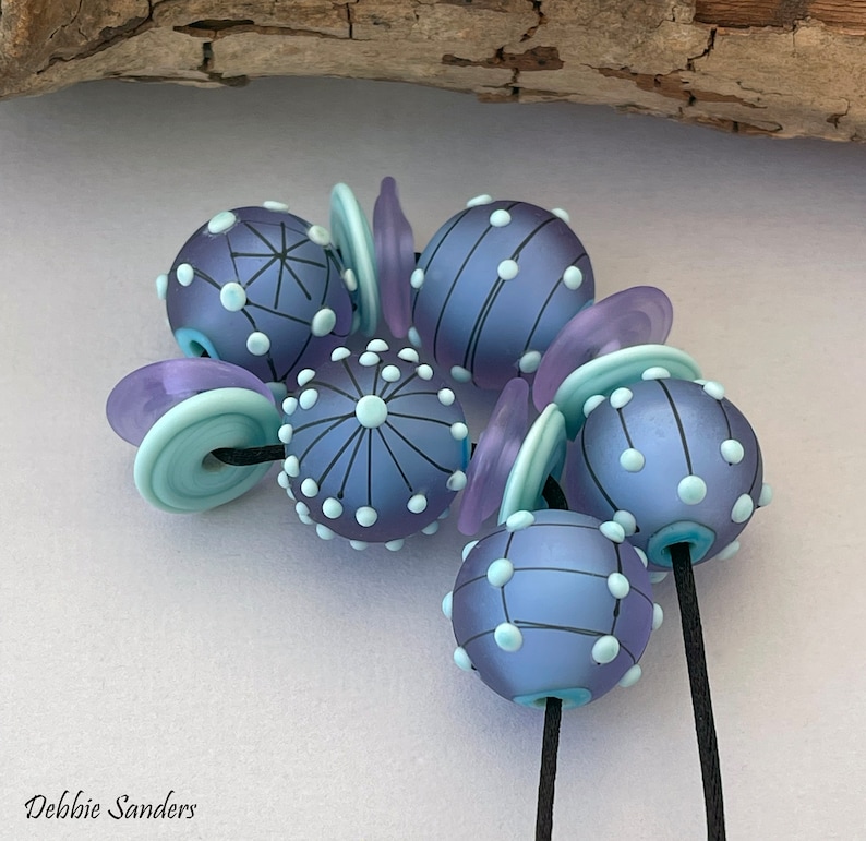 Pode incluir: Close-up de contas de vidro artesanais em tons de roxo e azul. As contas s&atilde;o decoradas com pontos brancos e linhas pretas. Algumas contas s&atilde;o redondas, outras planas ou cil&iacute;ndricas. As contas s&atilde;o enfiadas em um cord&atilde;o preto.