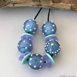 Pode incluir: Colar artesanal com contas de vidro lavanda fosco com desenhos de pontos e linhas brancas. Inclui espa&ccedil;adores azul claro e lavanda, enfiado em um cord&atilde;o preto. Design art&iacute;stico e &uacute;nico.