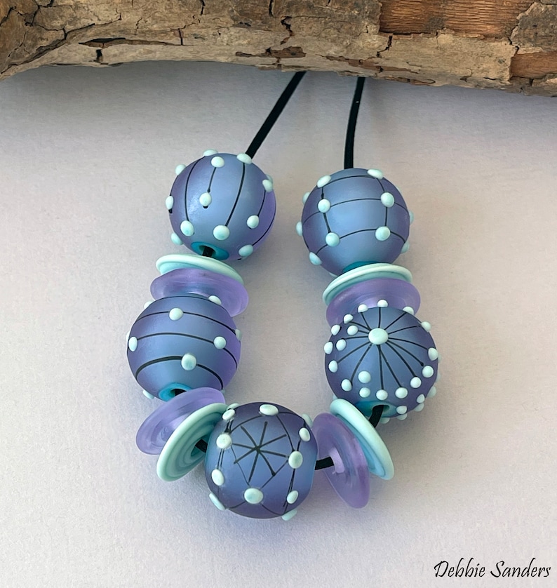 Pode incluir: Colar artesanal com contas de vidro lavanda fosco com desenhos de pontos e linhas brancas. Inclui espa&ccedil;adores azul claro e lavanda, enfiado em um cord&atilde;o preto. Design art&iacute;stico e &uacute;nico.