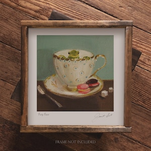May include: A green frog sits in a white teacup with blue polka dots. The teacup is on a white saucer with a spoon and two pink and brown macarons. The background is a light blue-green colour. The image is titled "Frog Face" and signed "Janet Hull".