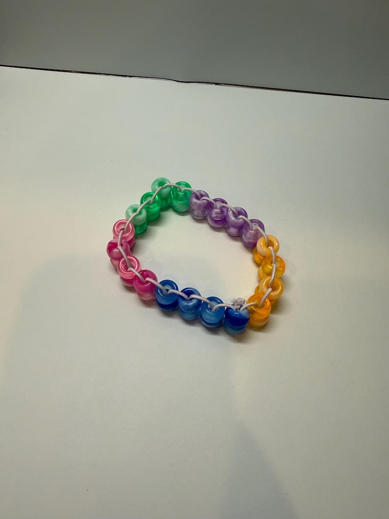 May include: A colorful bracelet made of round, translucent beads in a rainbow of colors: pink, green, blue, purple, and yellow. The beads are strung together on a white cord, creating a vibrant accessory.
