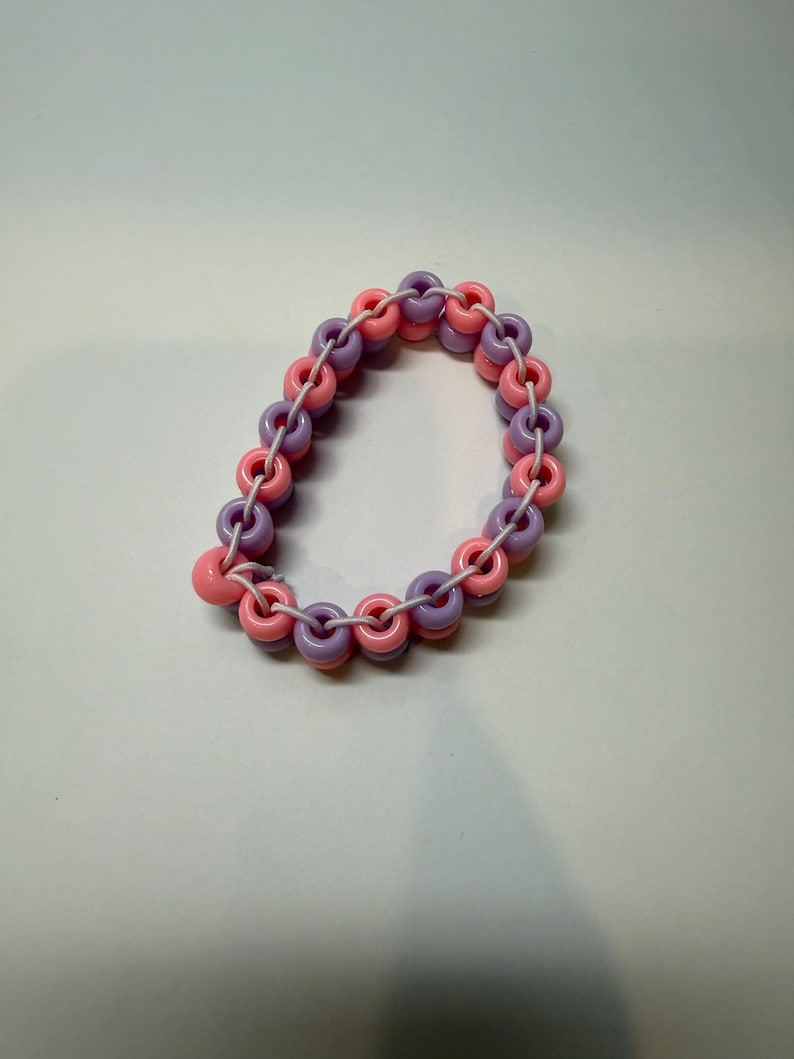 May include: A handmade bracelet featuring alternating pink and purple ring-shaped beads strung on a white cord. The bracelet has a playful, colorful design, perfect for adding a pop of color to any outfit. The beads are made of plastic.