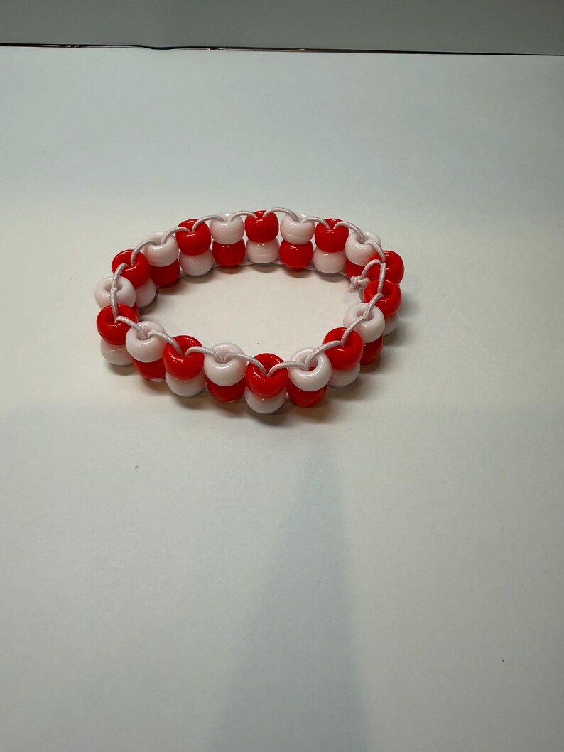 May include: A handmade bracelet featuring alternating red and white beads strung on a white cord. The beaded accessory is designed in a circular shape, suitable for wrist wear. The bracelet is a vibrant accessory.