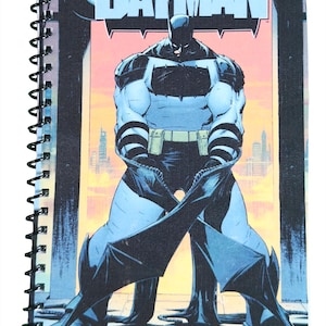 Puede incluir: Cuaderno con espiral con una portada de cómic. La portada presenta el texto "ABSOLUTE BATMAN" en azul y una ilustración de Batman en una pose dinámica. Un paisaje urbano es visible en el fondo. El cuaderno tiene una encuadernación en espiral negra.