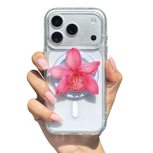 May include: Clear phone case with a vibrant pink orchid flower attached. The case has a silver camera module and a white MagSafe charger. The phone is held in a hand with manicured nails.