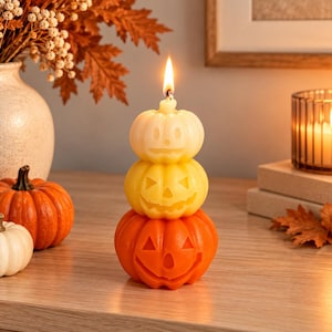 May include: A vertical stack of three carved pumpkin-shaped candles in a gradient of colors, starting with a pale cream top, a yellow middle, and an orange base. The top pumpkin has a lit wick, and each pumpkin features a carved jack-o'-lantern face.