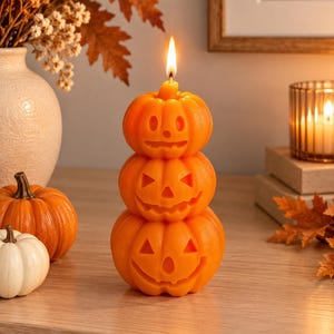May include: A stack of three orange wax candles shaped like pumpkins, each featuring a carved jack-o'-lantern face with a different expression. The top pumpkin has a lit wick, creating a flame at the peak. The pumpkins are stacked vertically.