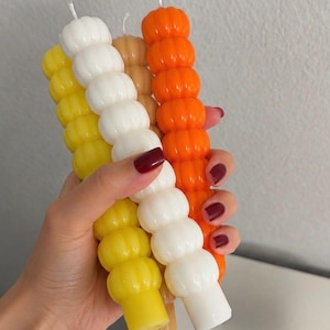 May include: A set of four long, cylindrical candles with a stacked bubble or sphere texture. The candles are colored in bright yellow, white, light tan, and vibrant orange. Each candle features a small, centered wick at the top end.