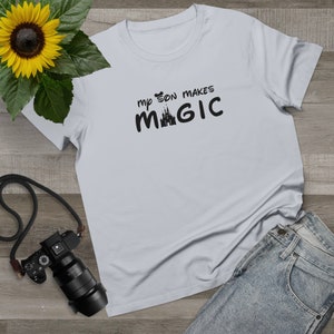 May include: A light gray t-shirt with the text "my son makes MAGIC" in black. The word "MAGIC" features a castle silhouette. The shirt is paired with blue jeans and a sunflower.