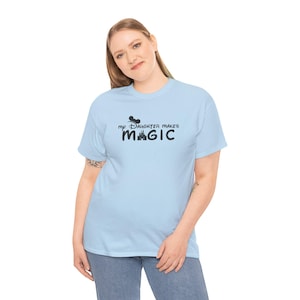 May include: Light blue t-shirt with the text "my Daughter makes MAGIC" in black. The design includes a silhouette of a mouse head and a castle. The shirt is made of a soft material.