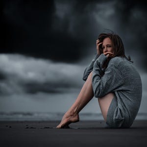 May include: A woman sits on a beach with her legs crossed, wearing a gray sweater and looking off to the side. The sky is dark and cloudy.