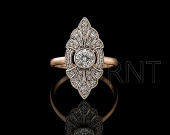 Puede incluir: Un anillo de oro rosa con un engaste ovalado alargado y puntiagudo, adornado con numerosas piedras pequeñas, redondas y transparentes, y una piedra central redonda más grande de talla brillante. Las letras RNT son visibles a la derecha.