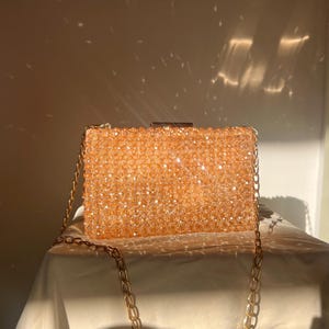 May include: A gold beaded clutch bag with a chain strap. The bag is made of small, sparkly beads and has a rectangular shape.