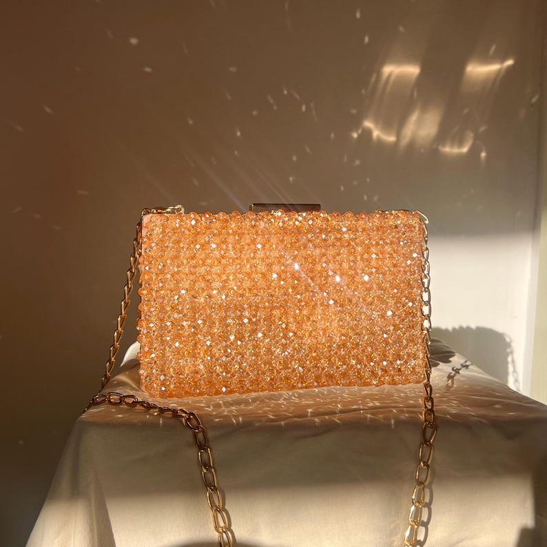 May include: A gold beaded clutch bag with a chain strap. The bag is made of small, sparkly beads and has a rectangular shape.
