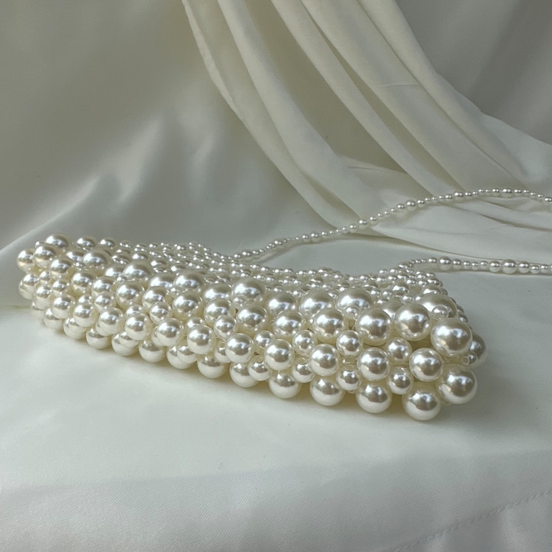 May include: A white, pearl-beaded clutch with a matching pearl strap. The clutch is rectangular and densely covered in round, off-white pearls. The background is a white, draped fabric.