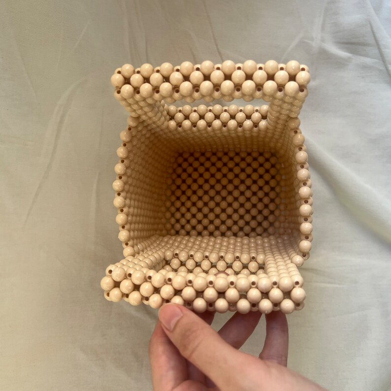 May include: A beige beaded handbag with a top handle. The bag is made of small, round beads that are woven together to create a square shape. The bag has a flap that can be closed with a button.