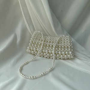 May include: A white beaded handbag with a pearl-like finish. The rectangular bag features a beaded strap and is set against a white, draped fabric background. The bag is a stylish accessory.