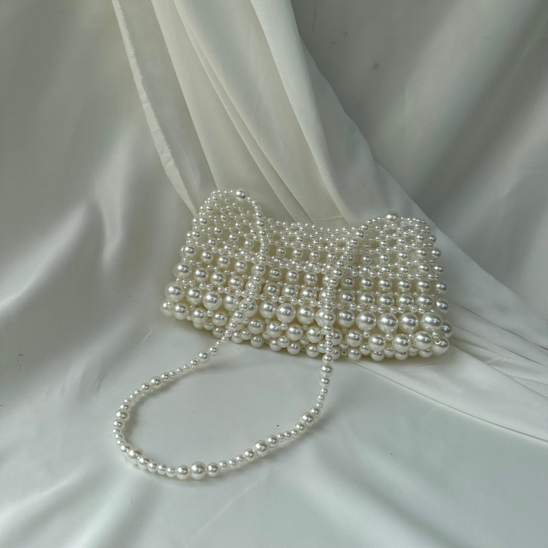 May include: A white beaded handbag with a pearl-like finish. The rectangular bag features a beaded strap and is set against a white, draped fabric background. The bag is a stylish accessory.