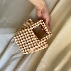 May include: A small, light beige, beaded handbag with a square shape and a top handle.