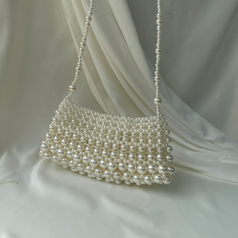 May include: A white beaded handbag with a shoulder strap. The rectangular bag is covered in small, round, white beads. The strap is also made of white beads. The bag is set against a white fabric background.