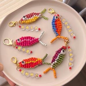 Puede incluir: Una colección de llaveros de peces de cuentas de colores. Cada pez tiene una combinación de colores diferente, incluyendo rojo, amarillo, rosa, azul, naranja y verde. Los llaveros tienen cierres dorados y etiquetas con nombres de cuentas como "Katy", "Bob", "Emma", "Isabel" y "Sara".