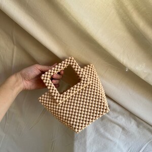 May include: A small, light beige handbag made of wooden beads. The bag has a square shape and a top handle.
