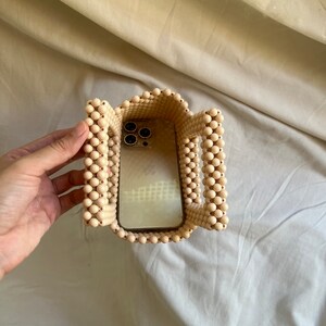 May include: A beige beaded bag with a phone inside. The bag is made of small, round beads and has a rectangular shape. The phone is a modern smartphone with a black camera lens.