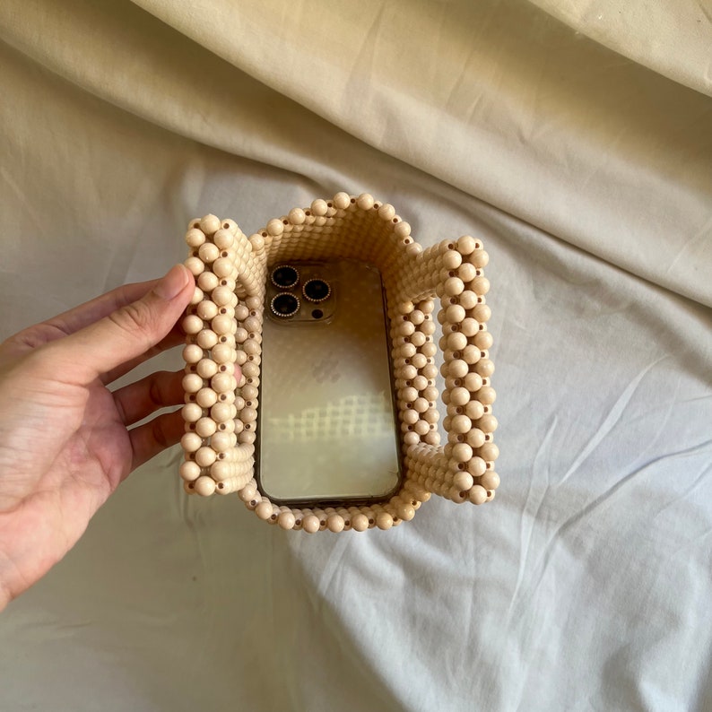 May include: A beige beaded bag with a phone inside. The bag is made of small, round beads and has a rectangular shape. The phone is a modern smartphone with a black camera lens.
