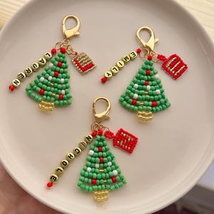 Può includere: Tre portachiavi a forma di albero di Natale con chiusure color oro. Ogni albero è fatto di perline verdi con accenti rossi e bianchi. Perline dorate scandiscono i nomi: LAUREN, EMILY e NICOLE. È attaccato un piccolo ciondolo a forma di scatola regalo rossa e oro.
