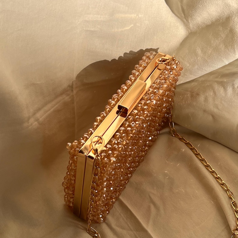 May include: A gold beaded clutch with a gold chain strap. The clutch is made of small, round beads that are sewn together to create a textured surface. The clutch has a gold clasp and a gold chain strap.