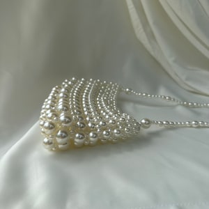 May include: A white, pearlescent beaded handbag with a curved, fan-like design. The bag is made of numerous small, round beads, with a beaded strap. The background is a white, draped fabric.