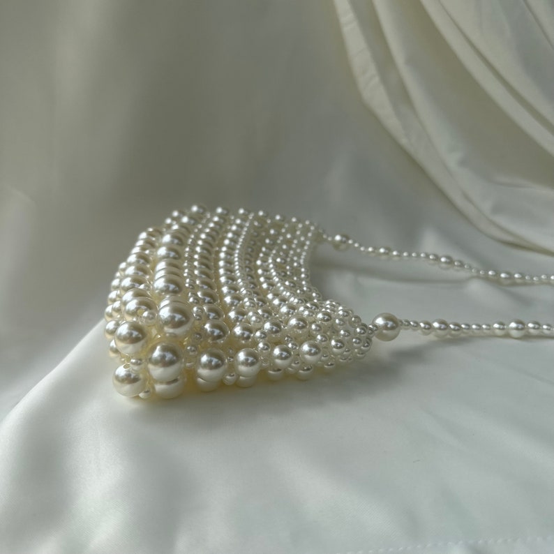 May include: A white, pearlescent beaded handbag with a curved, fan-like design. The bag is made of numerous small, round beads, with a beaded strap. The background is a white, draped fabric.