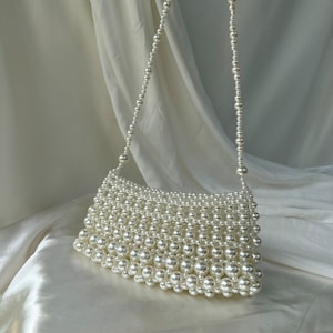 May include: A white beaded handbag with a shoulder strap. The bag is rectangular with a curved top edge, and the strap is made of smaller beads. The bag and strap are made of white, round beads.