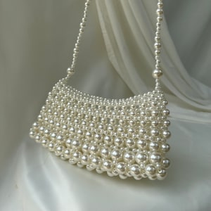 May include: Ivory-colored beaded handbag with a shoulder strap. The bag is made of small, round beads, creating a textured surface. The strap is also made of beads. The bag is set against a white, draped background.