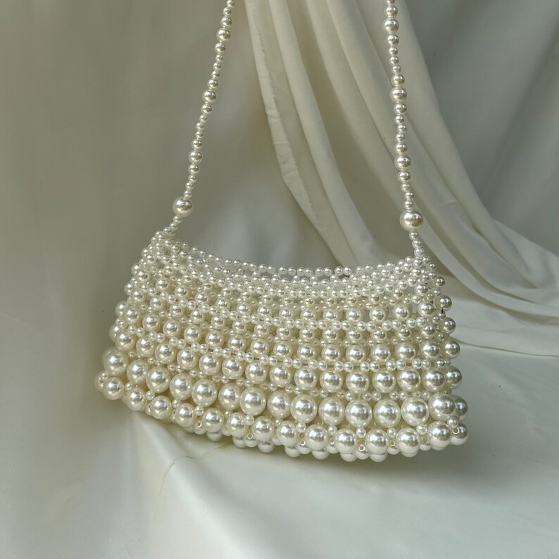 May include: A white beaded handbag with a shoulder strap. The bag is rectangular with a curved top and is made entirely of round, white beads. The strap is also made of white beads. The background is a white fabric.