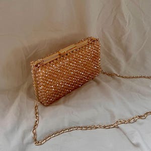 May include: A gold beaded clutch bag with a chain strap. The bag is made of small, round beads that are sewn together to create a textured surface. The chain strap is attached to the bag with two gold clasps.