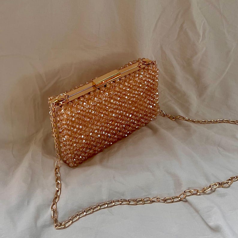 May include: A gold beaded clutch bag with a chain strap. The bag is made of small, round beads that are sewn together to create a textured surface. The chain strap is attached to the bag with two gold clasps.