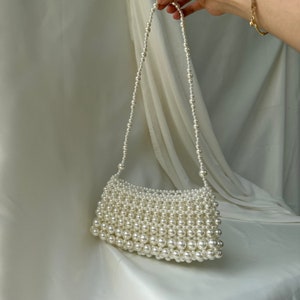 May include: A white, rectangular handbag made entirely of faux pearls. The bag features a shoulder strap also made of pearls. The pearls are a creamy white color and the bag is set against a white background.